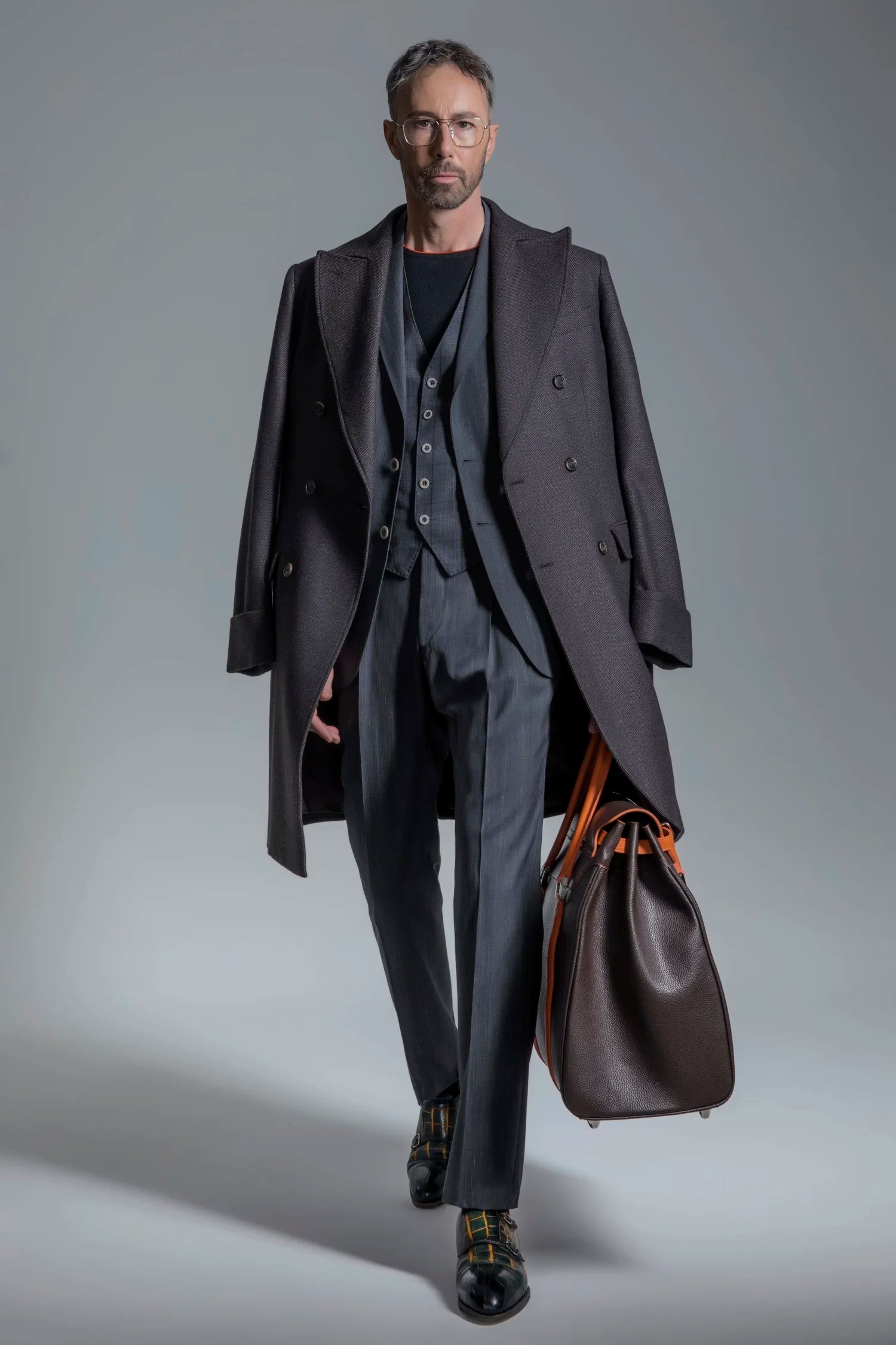 Man in a dark suit holding a brown leather bag against a gray background